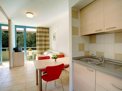 Salon lumineux avec coin repas, chaise rouge, kitchenette et accès au balcon avec vue sur les arbres.