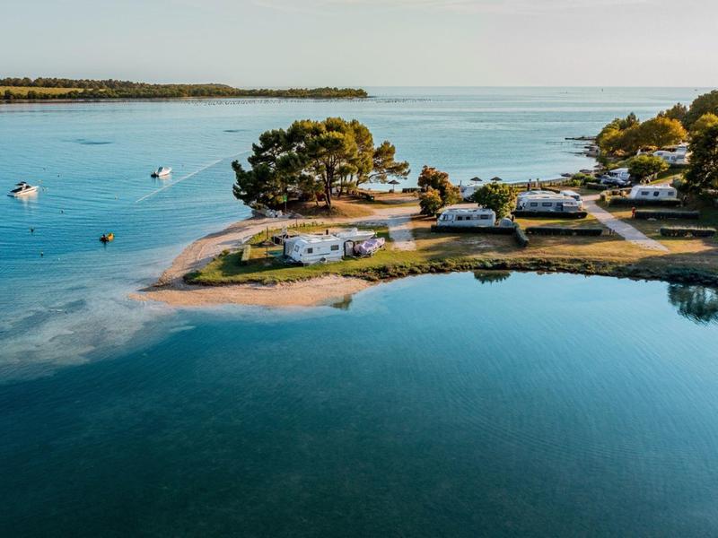 Camping côtier avec camping-cars, arbres et mer calme sous un ciel clair.