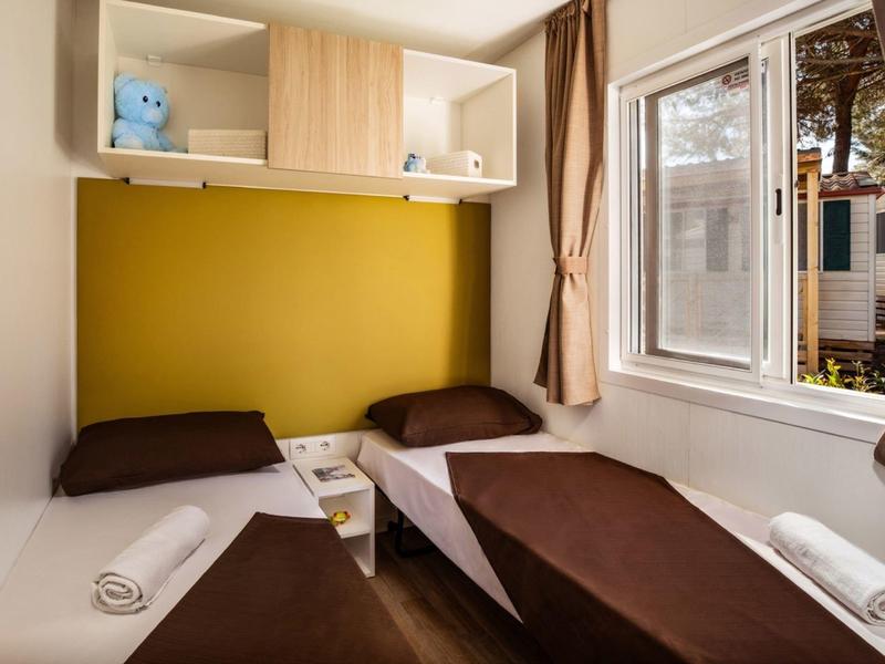 Zweibettzimmer mit braunen Betten, gelber Wand, großem Fenster und weißem Regal an der Wand.