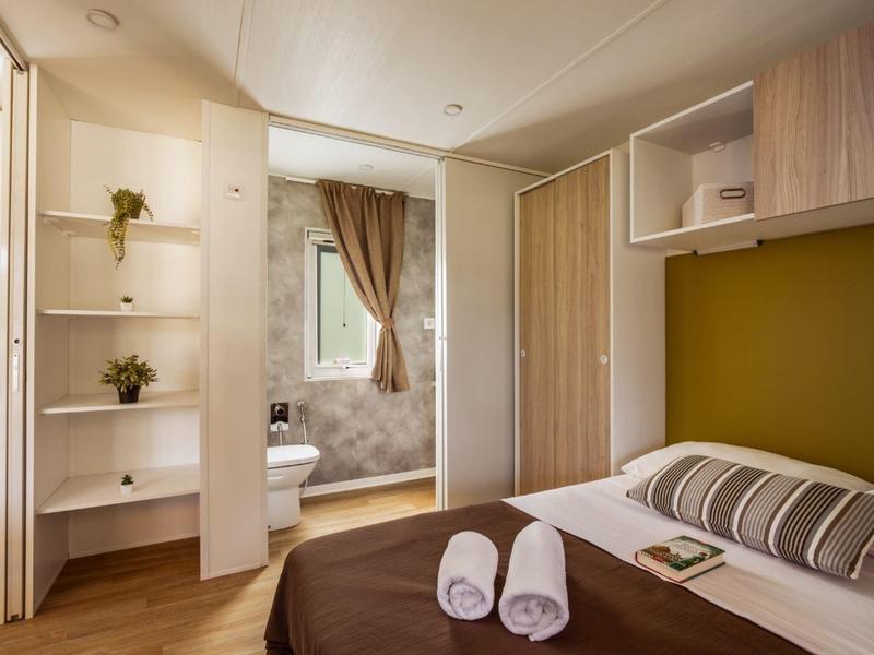 Helles Schlafzimmer mit Doppelbett, braun-weißer Bettwäsche, Regalen, Schrank und Fenster mit braunen Vorhängen.