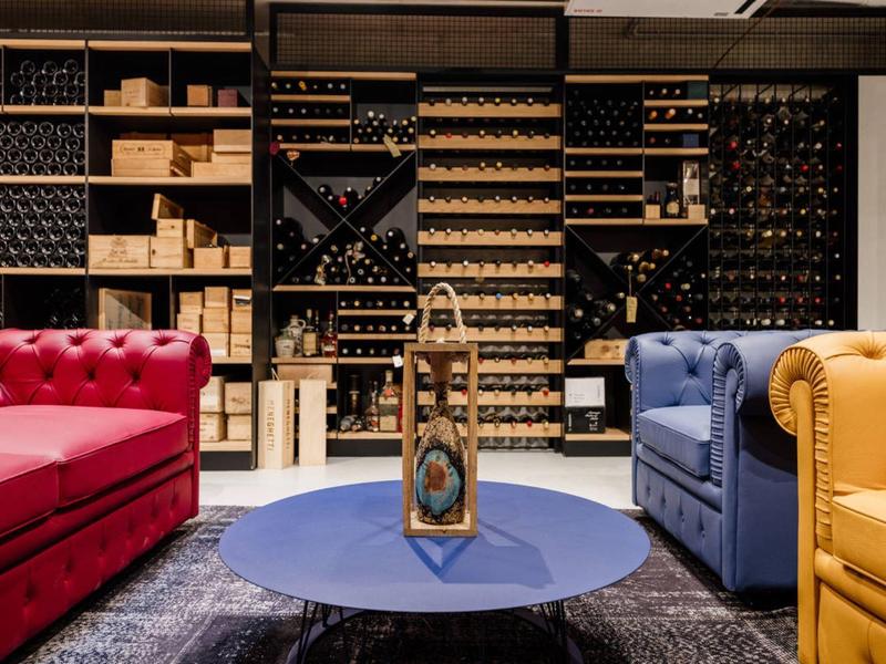 Przytulna część wypoczynkowa z kolorowymi sofami i regałami na wino w tle.