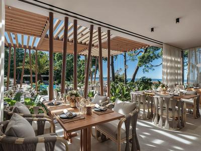 Moderní restaurace s dřevěným nábytkem s výhledem na bazén a bujnou zeleň.