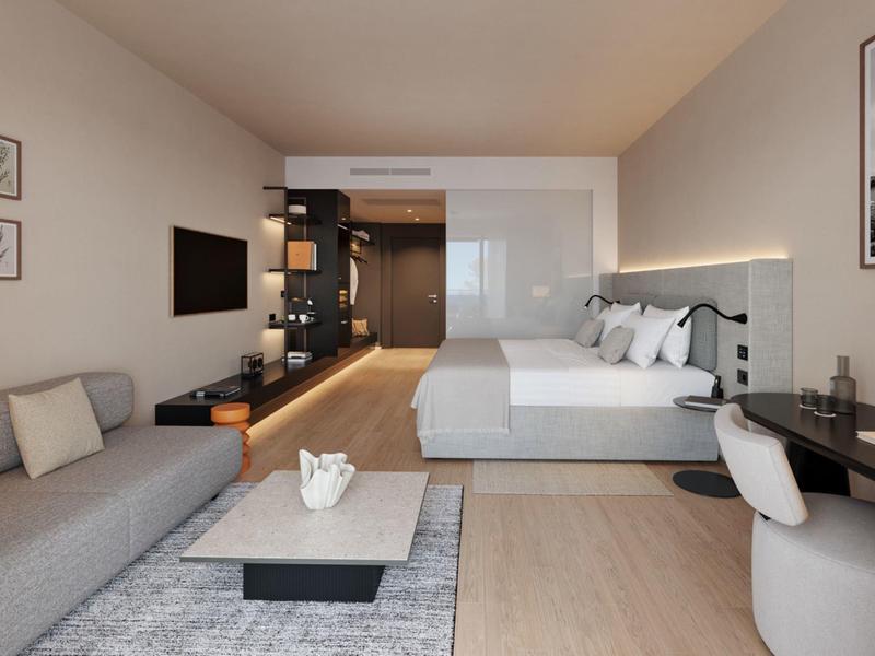 Modern furnished hotel room with double bed, sofa, TV, and desk in neutral tones.