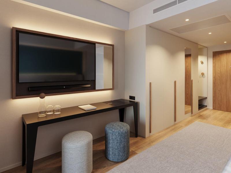Modern hotel room area with wall-mounted TV, console table, and two stools on carpeted floor.
