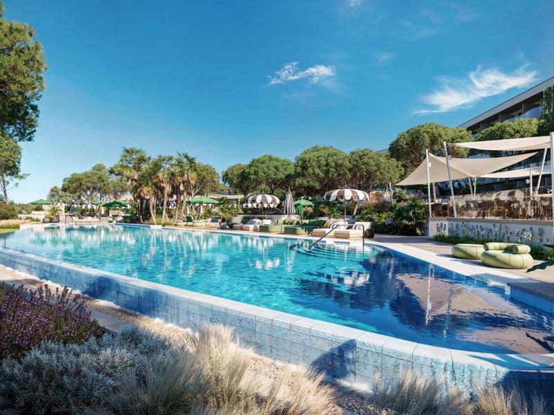 Modern hotelzwembad met parasols, ligstoelen en bomen onder een blauwe hemel.