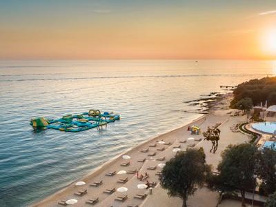 Strand mit Liegestühlen und Schwimmbereich bei Sonnenuntergang, neben Pool und Bäumen.