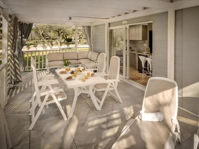 Terrasse mit Esstisch und weißen Stühlen, neben einem grauen Gebäude mit großen Fenstern.