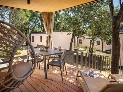 Balkon mit Holzboden, modernem Stuhl und Tisch mit sanften Vorhängen und Bungalows im Hintergrund.