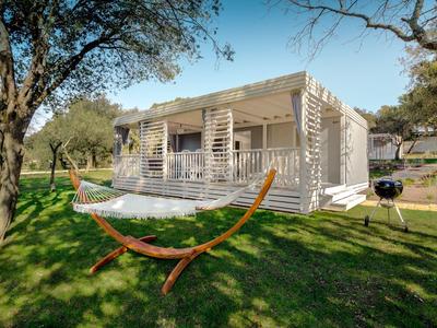Modernes Ferienhaus mit Terrasse, Hängematte und Grill auf grünem Rasen unter Bäumen.