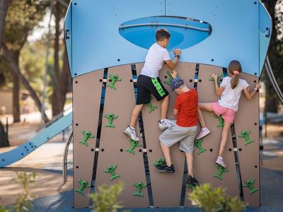 Drei Kinder klettern an einer Spielplatz-Wand im Freien bei sonnigem Wetter.