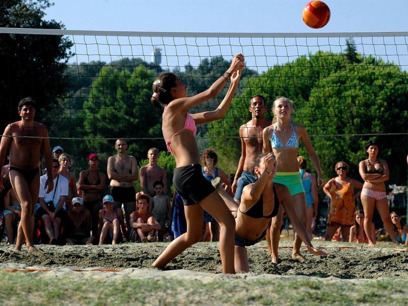 Mensen spelen volleybal op zand buiten met toeschouwers op de achtergrond.