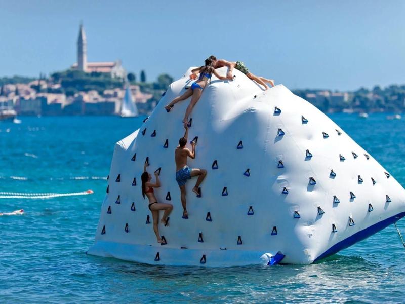 Mensen klimmen op een opblaasbare klimwand in het water met een kuststad op de achtergrond