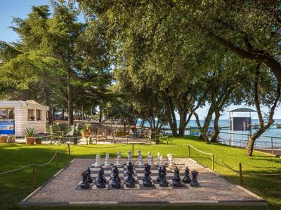 Duża plansza do szachów na trawie w ogrodzie hotelowym z widokiem na morze i drzewa.