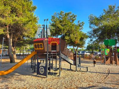 Plac zabaw na zewnątrz z okrętem pirackim, zjeżdżalniami i konstrukcjami do wspinaczki, otoczony drzewami.