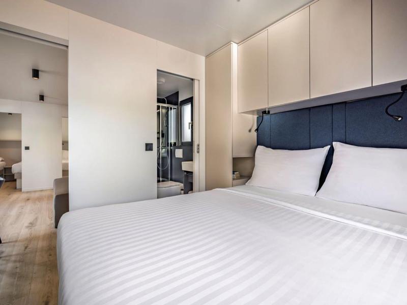 Nowoczesny pokój hotelowy z dużym łóżkiem, jasnymi ścianami i widoczną łazienką.