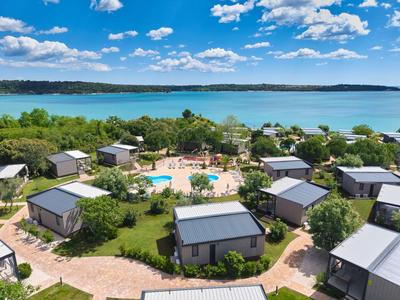 Holiday village with modern bungalows around a pool by a lake under a blue sky.