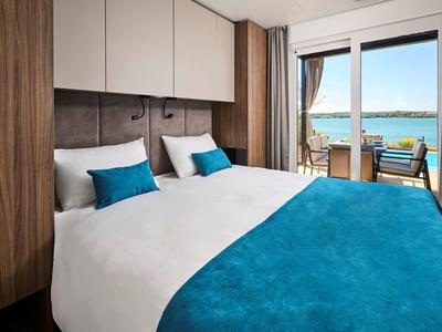 Nowoczesny pokój hotelowy z podwójnym łóżkiem, niebieską narzutą i tarasem z widokiem na wodę.