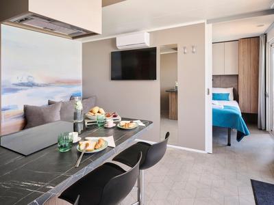 Nowoczesny pokój hotelowy z jadalnią, sofą, telewizorem i sypialnią w tle.