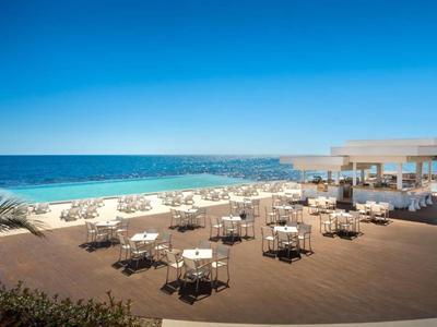 Open-air restaurant with tables and chairs by the beach under clear skies and calm sea.