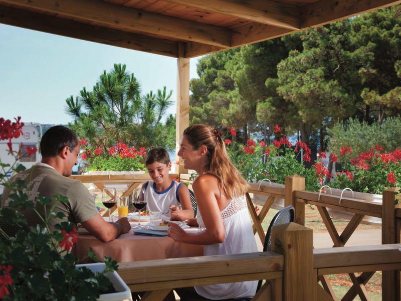 Rodzina siedzi na drewnianym tarasie hotelowym przy stole, otoczona kwiatami.