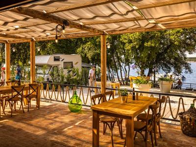 Zadaszony taras z drewnianymi krzesłami i stołami, widok na jezioro i drzewa w tle.