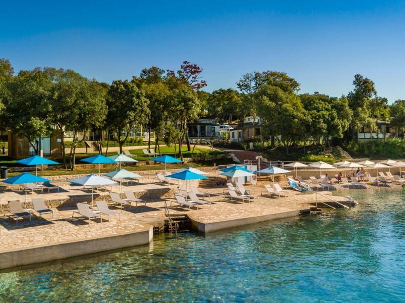 Obszar plaży z leżakami, niebieskimi parasolami i czystą wodą na tle zielonych drzew.