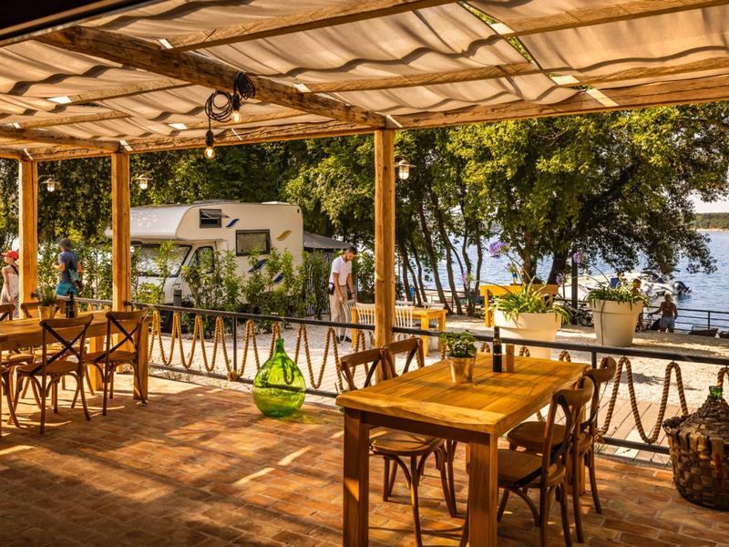 Zadaszony taras z drewnianymi krzesłami i stołami, widok na jezioro i drzewa w tle.