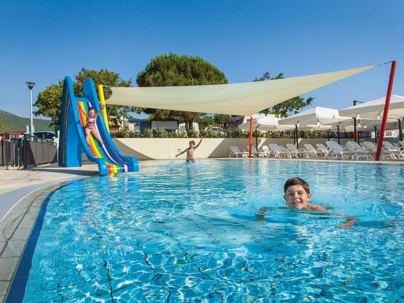 Niños nadan y se deslizan en la piscina de un parque vacacional con vela de sombra.
