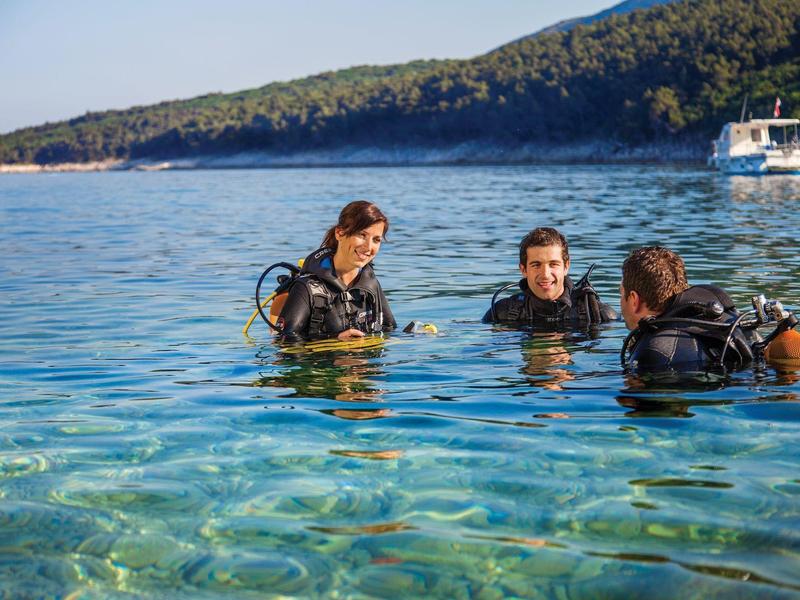 Tres buzos con equipo están en agua clara cerca de una costa boscosa.