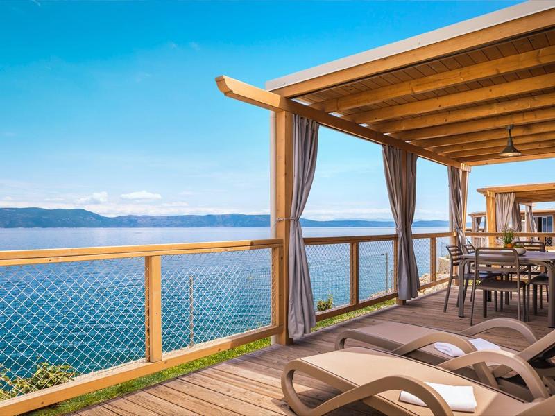 Balcón con tumbonas y mesa de comedor con vista a un lago y montañas al fondo.