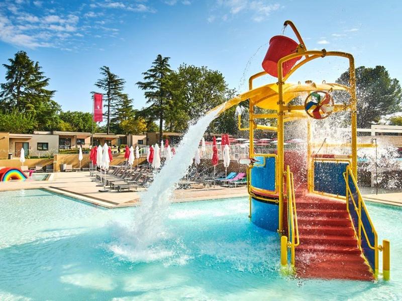 Parque acuático colorido con tobogán y agua salpicando en una piscina bajo un cielo azul.