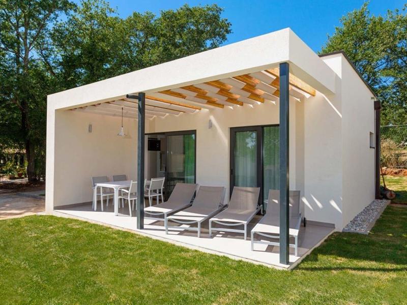 Pequeña casa de vacaciones moderna con terraza, tumbonas y mesa de comedor en entorno verde.