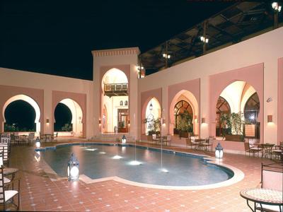 Innenhof mit großen Bögen, beleuchtetem Pool und Sitzbereichen in warmen Rosatönen bei Nacht.