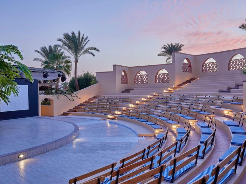 Anfiteatro al aire libre con filas curvas de asientos y palmeras al fondo al atardecer.