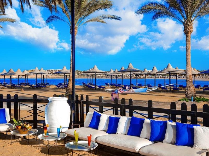 Terrazza con divano e vista sulla spiaggia con ombrelloni e palme sotto un cielo azzurro.