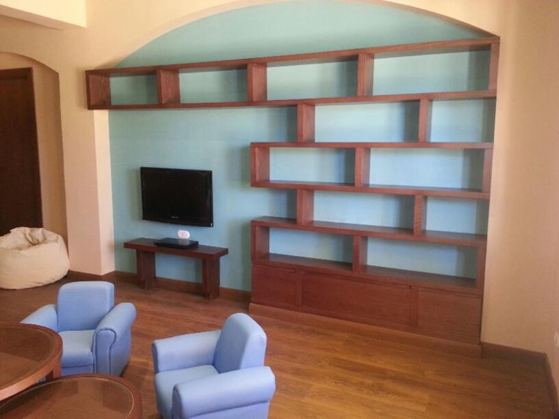 Salon avec étagères murales en bois, deux fauteuils bleus et un téléviseur sur meuble bas.