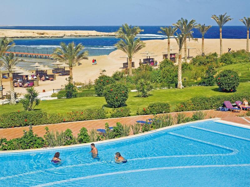 Piscina con nadadores y vista de palmeras, playa y mar al fondo.