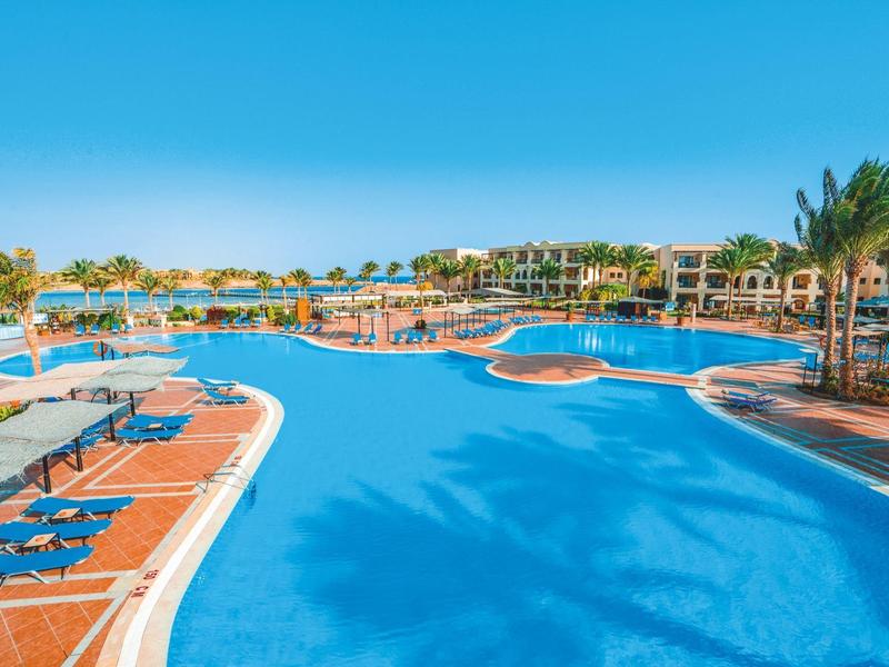Piscine spacieuse avec chaises longues et palmiers sous un ciel bleu clair.