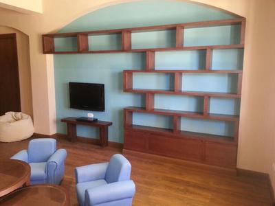 Salon avec étagères murales en bois, deux fauteuils bleus et un téléviseur sur meuble bas.