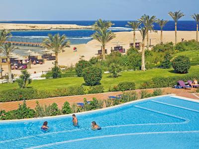 Piscina blu davanti a un giardino, spiaggia con palme e mare sullo sfondo.