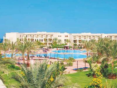 Gran complejo hotelero con piscina, palmeras y césped verde cuidado bajo un cielo azul.