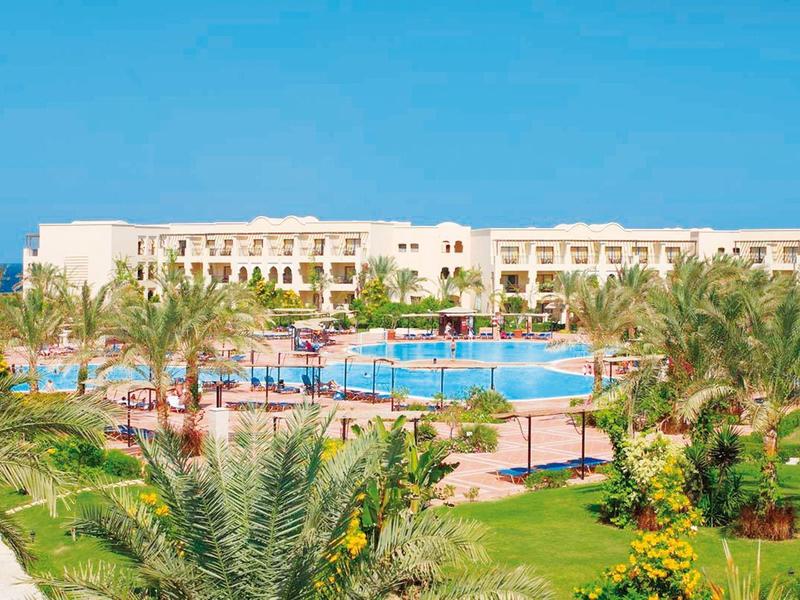 Gran complejo hotelero con piscina, rodeado de palmeras y césped bajo cielo azul.