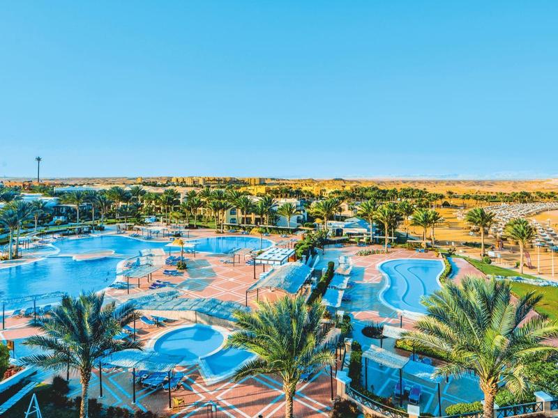 Panoramatický pohled na velký hotelový komplex s bazény, palmami a jasnou modrou oblohou.