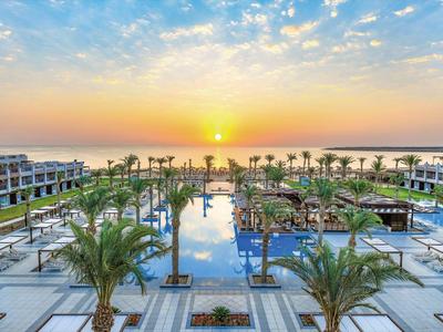 Luxe hotel met groot zwembad, palmbomen en zonsondergang boven de zee op de achtergrond.