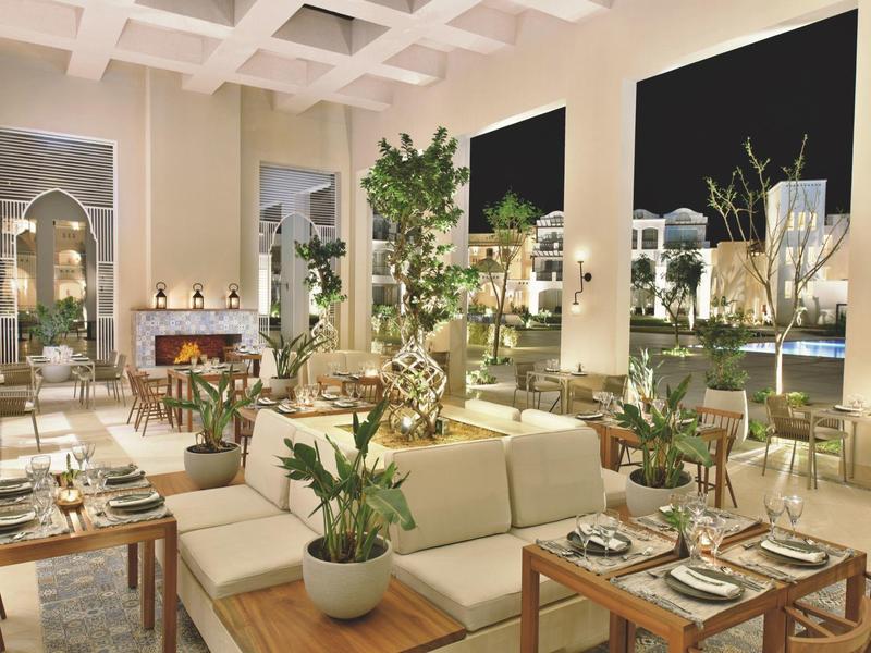 Salotto d'hotel elegante con divani bianchi, tavoli apparecchiati e vista notturna sul giardino.