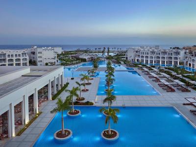 Grande piscina dell'hotel con palme e lettini con vista sul mare al tramonto.