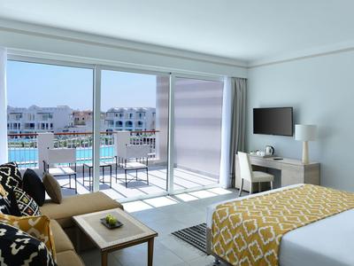 Bright hotel room with bed, sofa, desk, and balcony overlooking a resort pool and buildings.