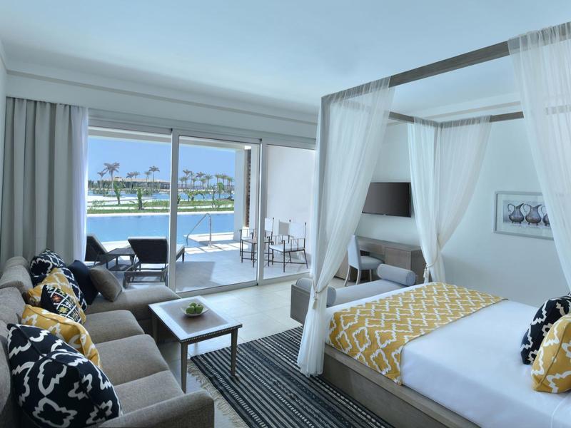 Bright hotel room with a canopy bed, sofa, and a balcony overlooking water and palm trees.