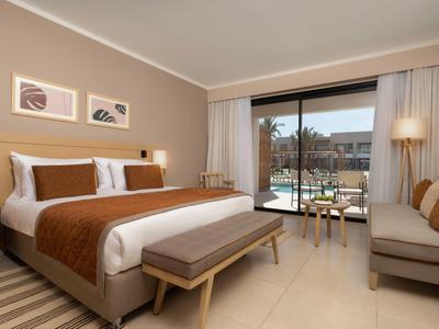 Modernes Hotelzimmer mit großem Bett, Sofa, Tisch und Blick auf einen Pool durch große Fenster.