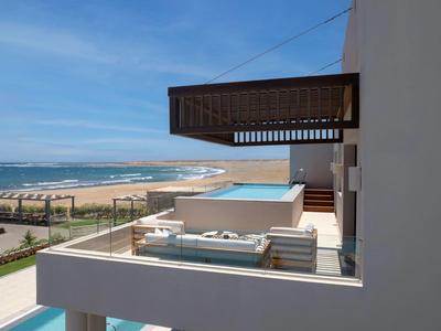 Balcone moderno con divano che si affaccia su piscina e spiaggia infinita con cielo azzurro.